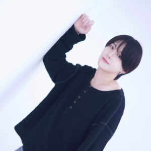 Portrait of a person with short black hair wearing a dark top, leaning against a white wall, facing slightly sideways.