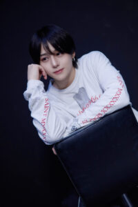 Person with short black hair rests their chin on a hand, wearing a white long-sleeve shirt and holding a large black bag against a dark background.