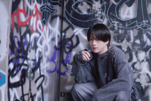 Young person with short black hair in a ripped gray denim jacket sits against a graffiti-covered wall.
