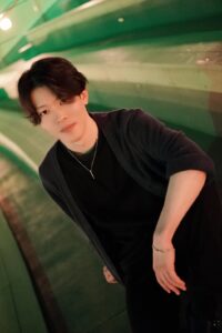 Young man with dark hair wearing a black shirt and cardigan, leaning forward in a green-lit indoor space.
