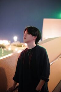 Young man with dark hair in a black shirt and cardigan stands on an outdoor staircase at night, city lights blurred behind him.