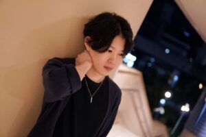 Young man leaning against a beige wall at night, hand to neck, wearing a black shirt and dark cardigan with a pendant necklace.
