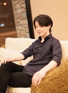 Young man in a dark shirt lounging on a cream sofa with a woven chair nearby.