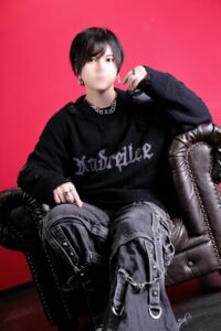 Person with short black hair and a blurred face sits in a dark brown leather chair against a bright red background, wearing a black distressed sweater and silver jewelry.