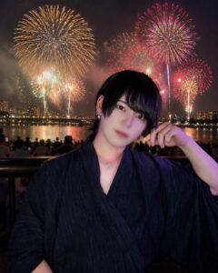 Person posing in a dark kimono-style outfit with a hand near their head, fireworks bursting in the night sky over a river and city lights in the background.