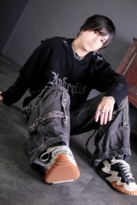 Person sitting on the floor in a black distressed sweater, gray cargo jeans, and chunky sneakers, with short dark hair and a silver chain, face blurred for privacy.