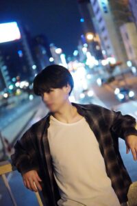 Young man posing on a city street at night, wearing a white t‑shirt and a plaid shirt, leaning with hands on a railing amid colorful bokeh lights.