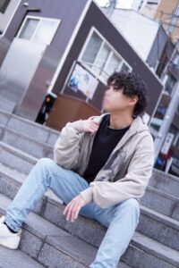 Young man with curly hair sits on urban concrete stairs, wearing a beige hoodie, black shirt, light jeans, looking to the side.