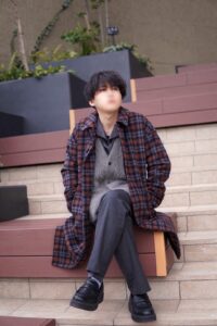 Young man sits on outdoor steps wearing a red plaid overcoat, gray cardigan, and dark trousers, face blurred for privacy.