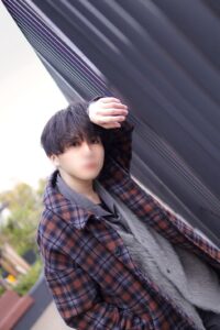 Outdoors portrait of a person in a brown plaid coat and gray sweater, leaning against a dark diagonal wall with their hand resting on their head; face blurred for privacy.