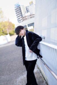Young man in a white cable-knit sweater and black coat leaning on a railing in an urban outdoor setting.