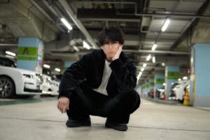 Person crouching in a dim indoor parking garage, hand resting on cheek, wearing a black outfit and glasses.”,