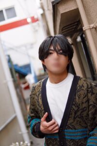 Person standing in a narrow urban alley wearing a beige and black patterned cardigan over a white T‑shirt, looking at the camera.