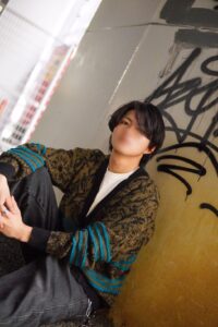 Young person with black hair sits against a graffiti-covered wall in an urban alley, wearing a patterned cardigan with teal stripes and a white shirt, looking toward the camera with a blurred face.