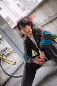 Young person with dark hair sits on a metal platform in an urban alley, hands clasped, yellow bicycle in the background