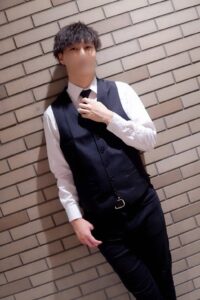 Young man in a white shirt and black vest adjusts his tie while leaning against a beige brick wall.