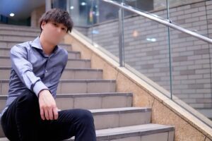 Man sitting on indoor stairs in a light blue shirt and dark pants, looking at the camera. (shot in a modern building with glass railing)