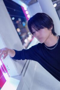 Young person with black hair and a silver chain necklace leans on a white ledge in a neon-lit, blue-purple night scene.