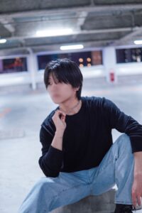 Person sitting on a concrete ledge in a dim underground parking garage, wearing a black shirt and light jeans, hand resting near the neck with a blurred face.