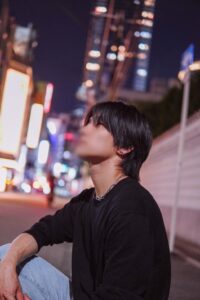 Young man with black hair wearing a black long-sleeve shirt and silver chain necklace sits on a city sidewalk at night, blurred neon lights in the background.]}}{