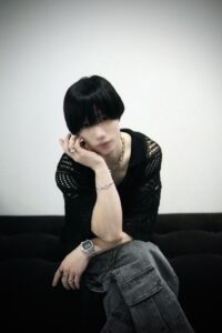 Person with short black hair posing on a dark couch, resting chin on hand, wearing a black knit top, silver jewelry, and gray cargo jeans against a plain wall.