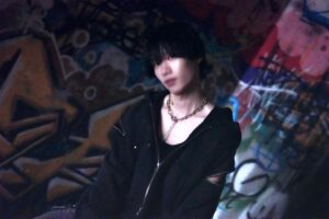 Portrait of a person with short black hair in a dark jacket and chunky chain, posing in a graffiti-filled alley.