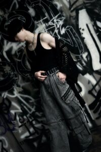 Person in a black tank top and gray cargo pants stands against a graffiti-covered wall, head tilted downward.