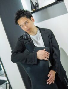 Young man in a black blazer hugs a black dress form against a modern interior backdrop.