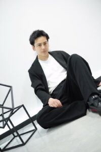 Young man in a black suit and white shirt sits on the floor against a white wall, with abstract black metal frames nearby.