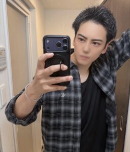 Young man with short black hair wearing a plaid shirt, taking a mirror selfie with a black iPhone in a bathroom.
