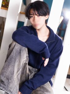 Portrait of a young person with dark hair in a navy sweater and gray jeans, leaning against white panels.
