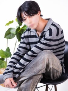 Person seated on a black chair, wearing a gray and black striped sweater and faded jeans with a green plant in the background.